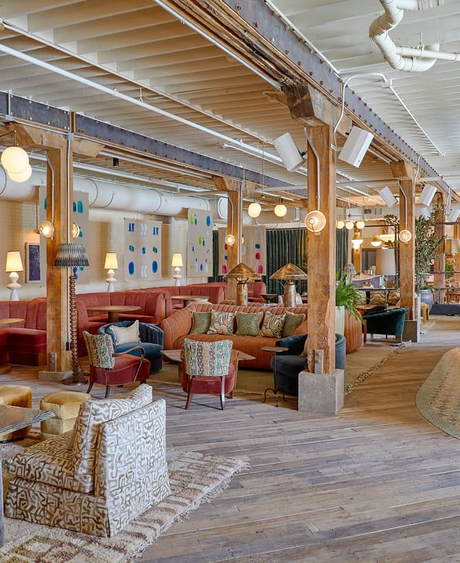 A spacious lounge with eclectic seating, including patterned chairs and sofas, is situated in an industrial-style building. The setting includes warm lighting, colorful wall art, and a bar area.