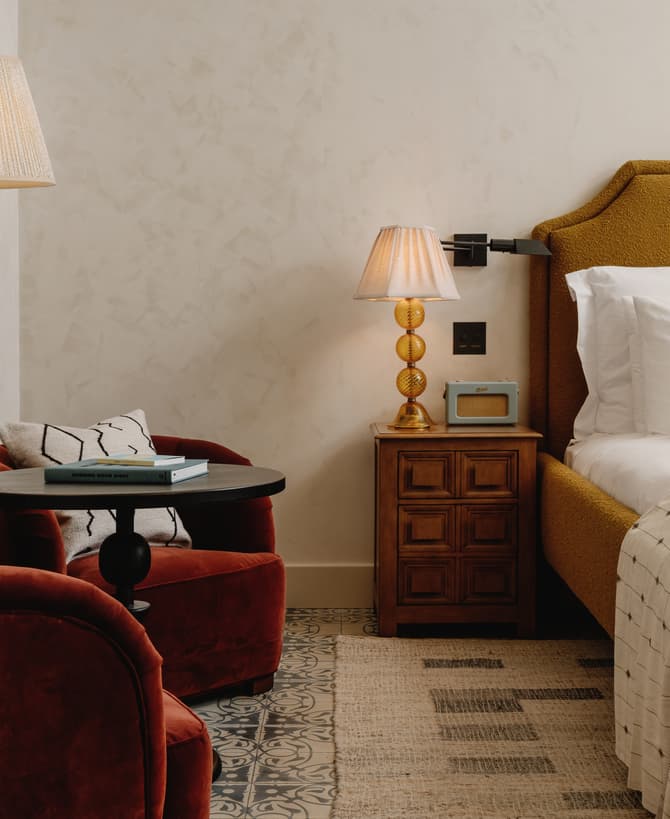 A cozy bedroom features a mustard yellow bed with white linens and a patterned pillow, flanked by a wooden nightstand with a lamp and radio. Two red armchairs, a round table with books, and a floor lamp complete the setting.