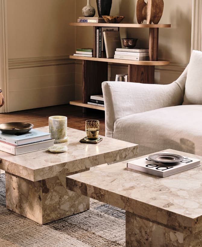 Marble coffee tables displaying books and decor sit between a beige sofa and a brown leather chair; background includes wooden shelves with books and decorative items. Visible book title: "ATHENS RIVIERA".