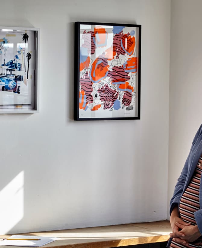 A woman with long hair, wearing a blue coat, stands gazing at two framed artworks on a white wall in a sunlit gallery space.