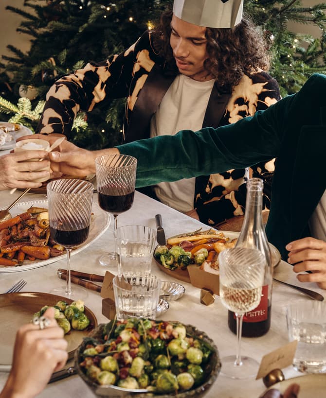 People are seated at a festive dining table, sharing food and drinks, surrounded by decorated Christmas trees and lit candles. Various dishes and wine glasses are spread across a white tablecloth.