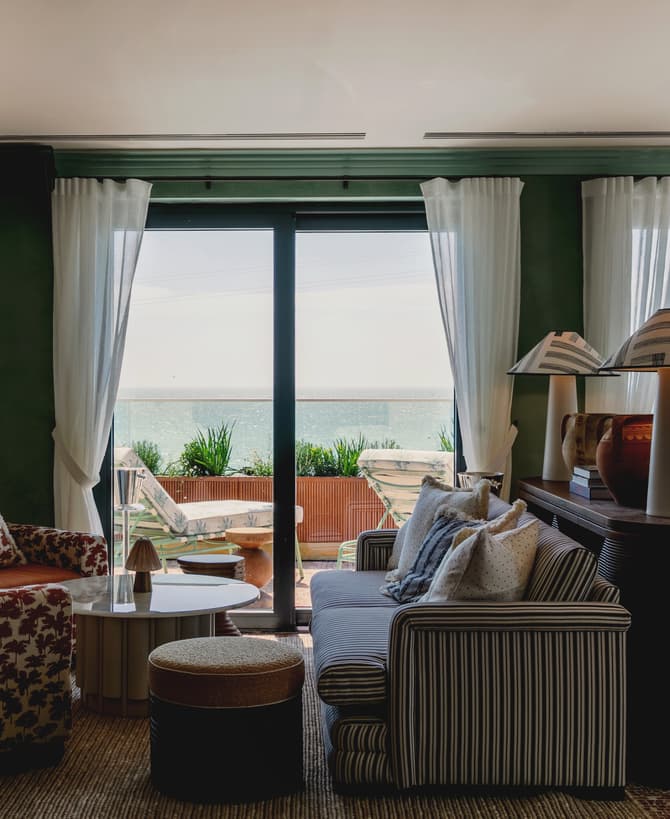 A brightly lit lounge with patterned armchairs, a striped couch, and two table lamps. Floor-to-ceiling windows with sheer curtains overlook an outdoor patio and ocean view.