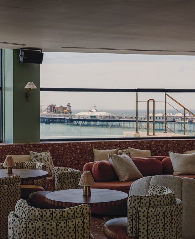 Round tables and cushioned seating face large windows overlooking a seaside pier. The interior features patterned armchairs, sofas, and small lamp fixtures, with views of the ocean and pier structures.