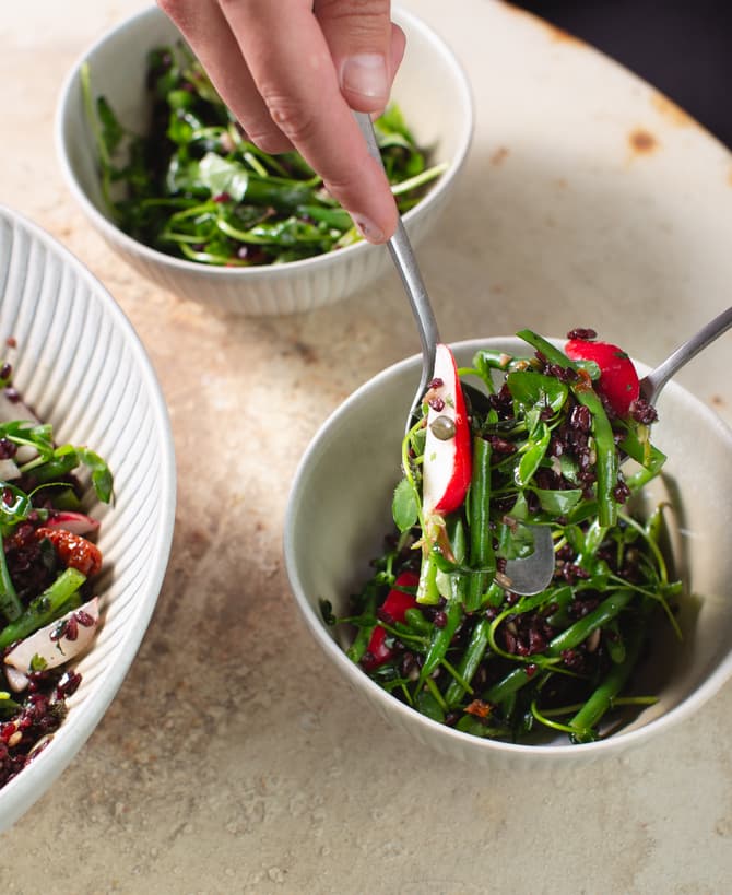 Hands scoop mixed green salad with radishes and black rice from a large bowl into a smaller one on a light, textured tabletop.