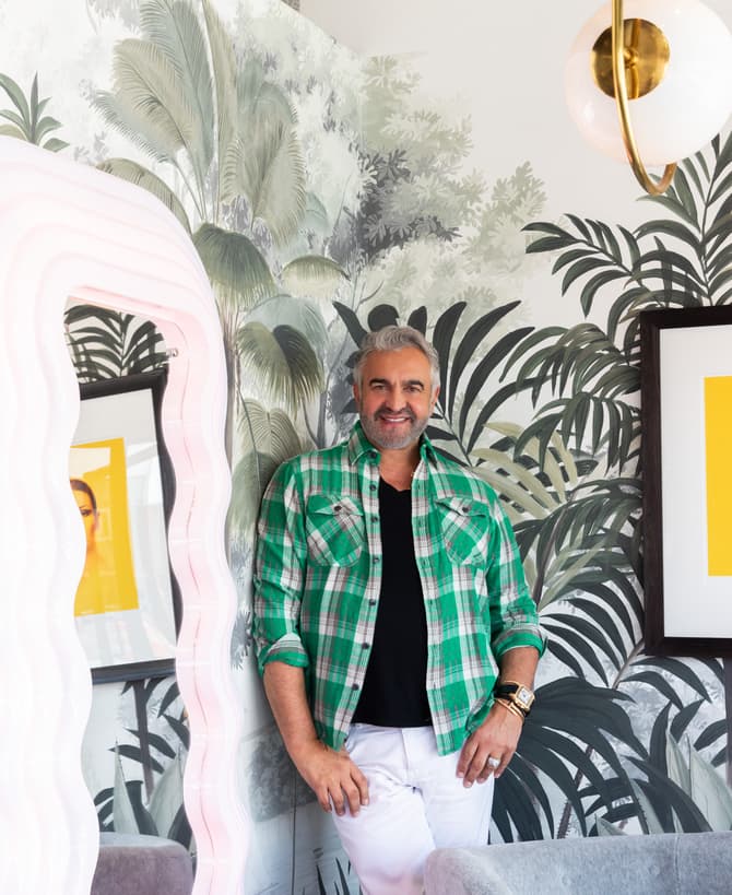 A man in a green plaid shirt and white pants stands by a wavy mirror, in a room with tropical-themed wallpaper and modern wall art.