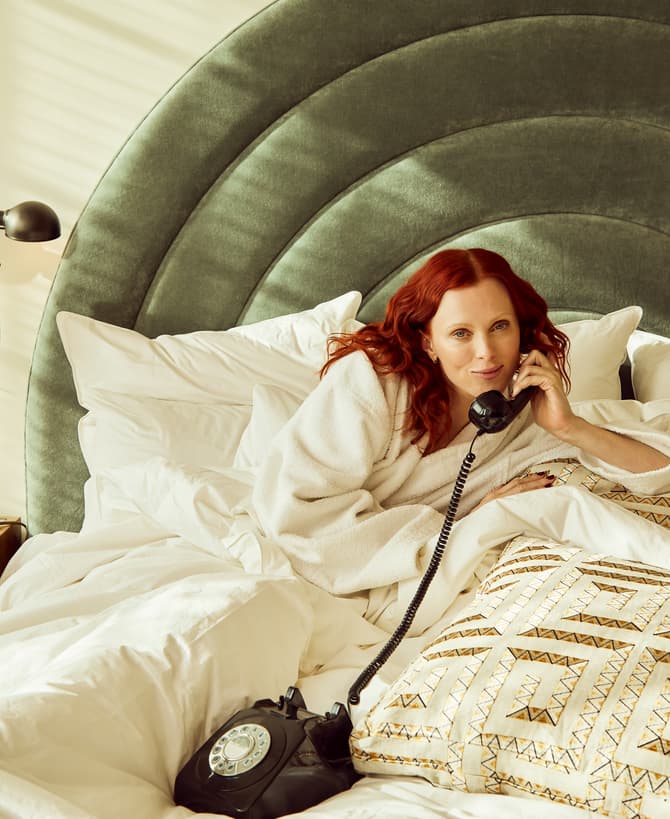 A woman, wearing a white bathrobe, lies on a bed talking on a rotary phone. The bed is adorned with white bedding and patterned pillows; a lamp sits on a wooden bedside table.