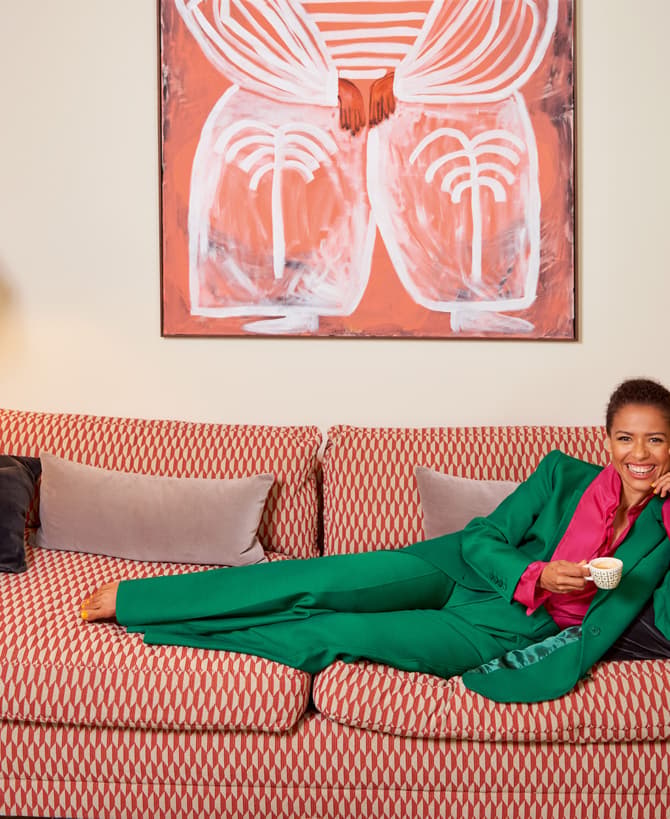 Woman in a green suit lounges on a patterned couch, holding a teacup, smiling; contemporary living room setting with a lamp on the left and abstract art on the wall.