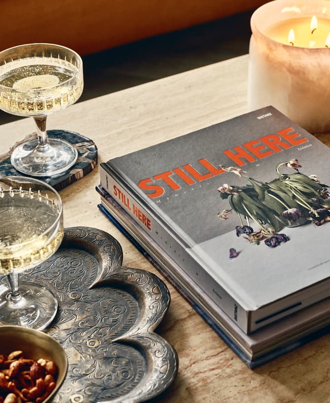 Two champagne flutes on coasters, a lit candle, a brass bowl of nuts, and books titled "STILL HERE" rest on a carved metal tray and a wooden table.