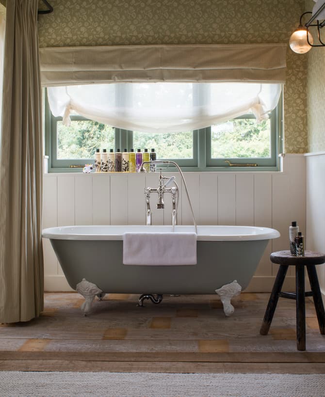 White clawfoot bathtub with a white towel draped over it, situated in a vintage bathroom. Surroundings include a wooden stool holding toiletries, a radiator, large window with a view of greenery.