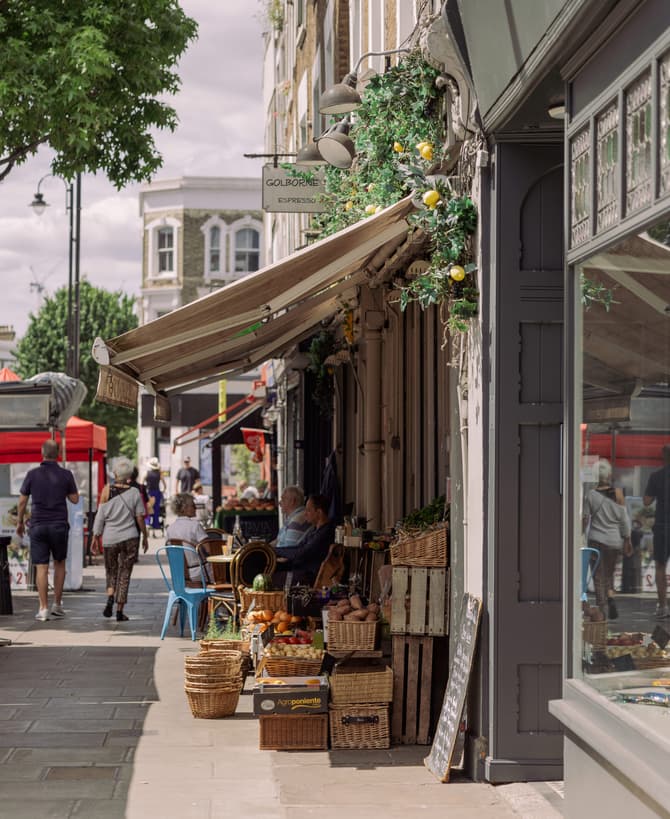 A street market featuring outdoor cafes with people seated, fruits and vegetables displayed in baskets. Passersby walk along the sidewalk. Text: "GOLBORNE ESPRESSO" on a shop sign.