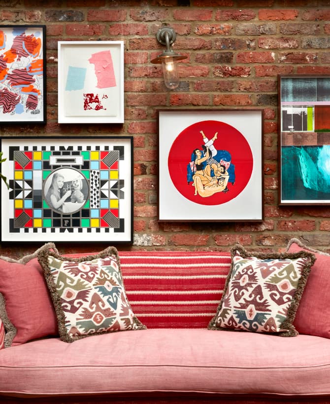 A red couch with patterned pillows sits against a brick wall displaying various framed artworks and a potted plant to the left.