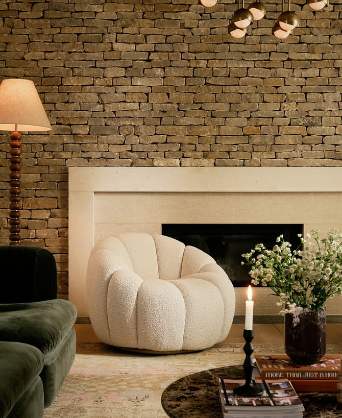 A living room features a green velvet sofa with cushions, two white plush armchairs, a stone fireplace, a standing lamp, and a marble coffee table with a candle, books, and a vase of flowers.