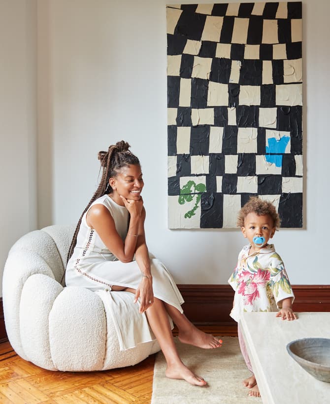 A woman sits on a cushioned chair, smiling, while a toddler with a pacifier stands nearby; they are in a cozy, light-filled room with wooden floors, near a modern art painting.