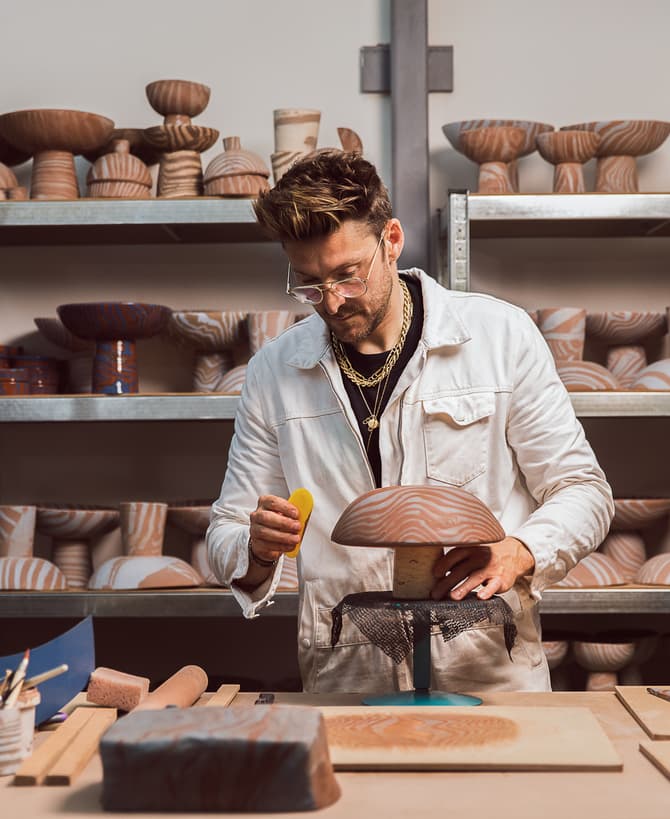 A person meticulously handcrafts a wooden bowl, using a tool for detailing, in a workshop surrounded by various shaped and patterned wooden bowls on metal shelves and a cluttered workbench.