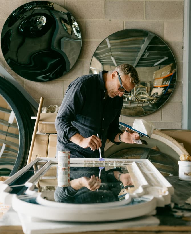 A craftsman paints a frame in a workshop with circular mirrors mounted on brick walls, surrounded by tools, boxes, and materials.
