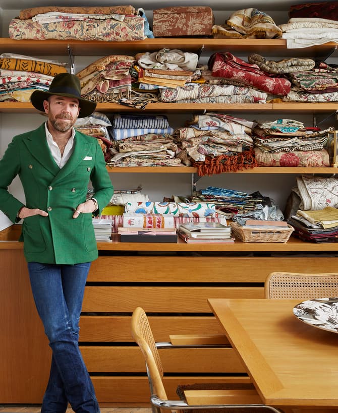 A man stands with hands in pockets, wearing a green blazer and hat, in front of shelves filled with folded fabrics and books, beside a wooden table with a decorative dish.