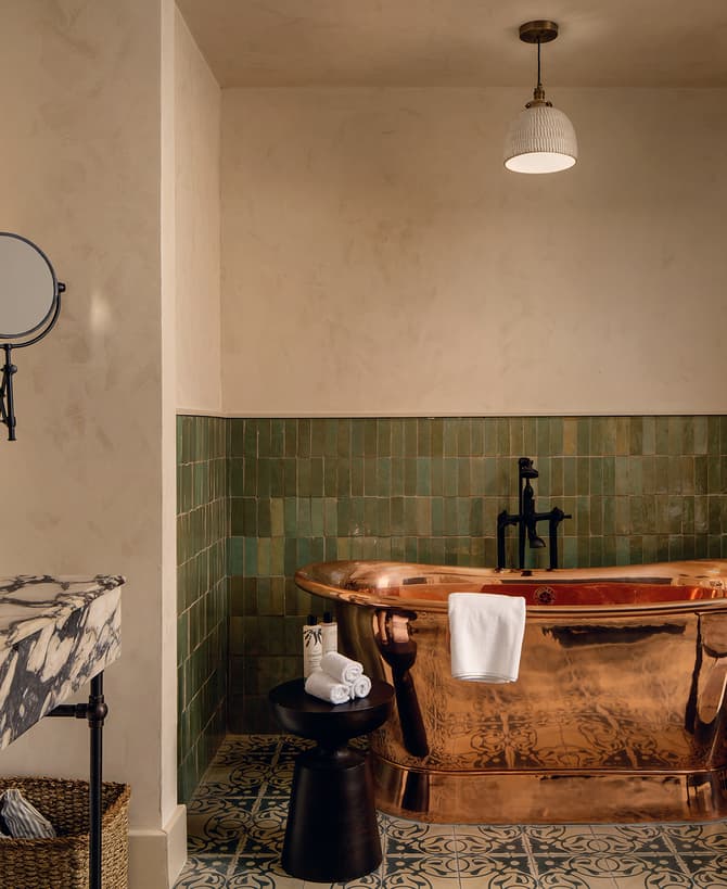 Copper bathtub with a towel hanging over it, set against green tiled walls. Marble sink with products, mirror, and magnifying mirror. Black stool with rolled towels and a plant.