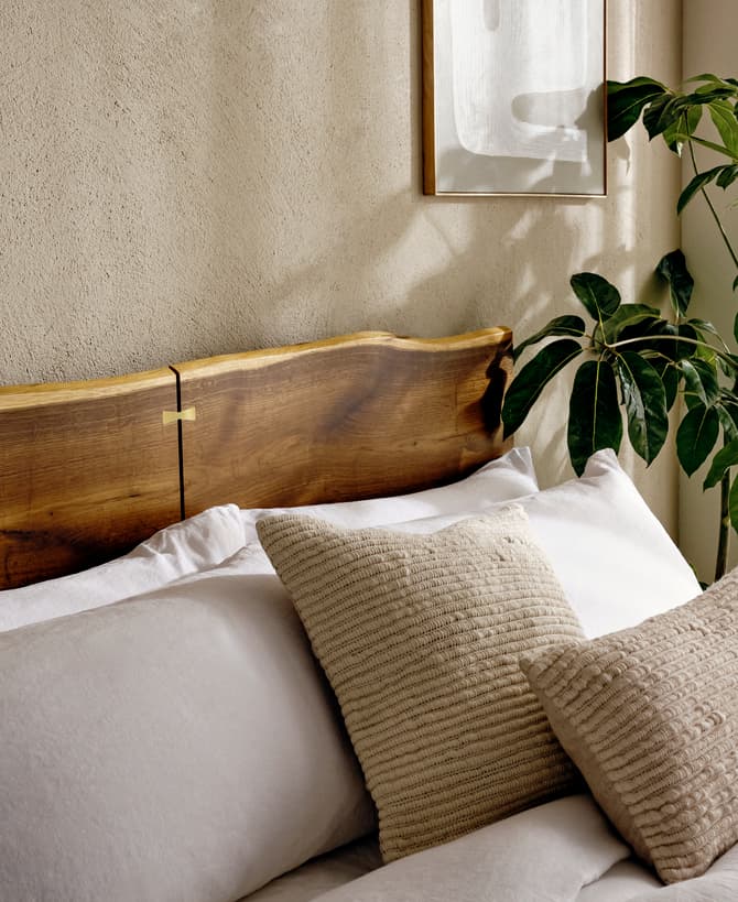 A cozy bed features a wooden headboard and cream-colored pillows; next to it is a potted leafy plant, under soft natural light from a window with sheer curtains.