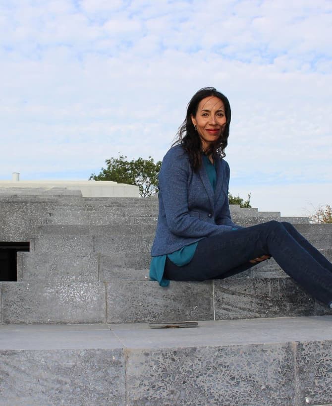 A woman wearing a blue jacket and jeans sits on concrete steps of a modern, grey stone structure, with a partly cloudy sky in the background.