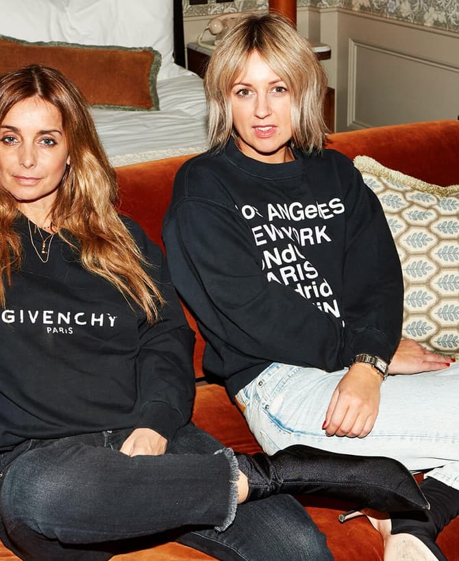 Two women sit relaxed on an orange couch. One wears a "Givenchy Paris" sweatshirt, while the other wears a sweatshirt listing city names. Background includes a bed and a bar cart.