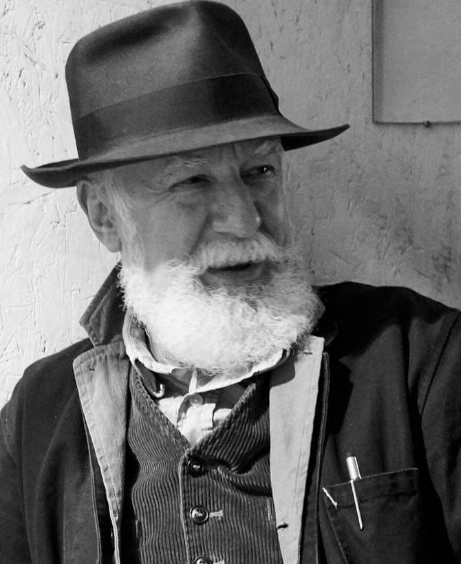 Bearded man wearing a hat and jacket sits against a textured wall, smiling slightly, with a pen visible in his jacket pocket in a dimly lit room.