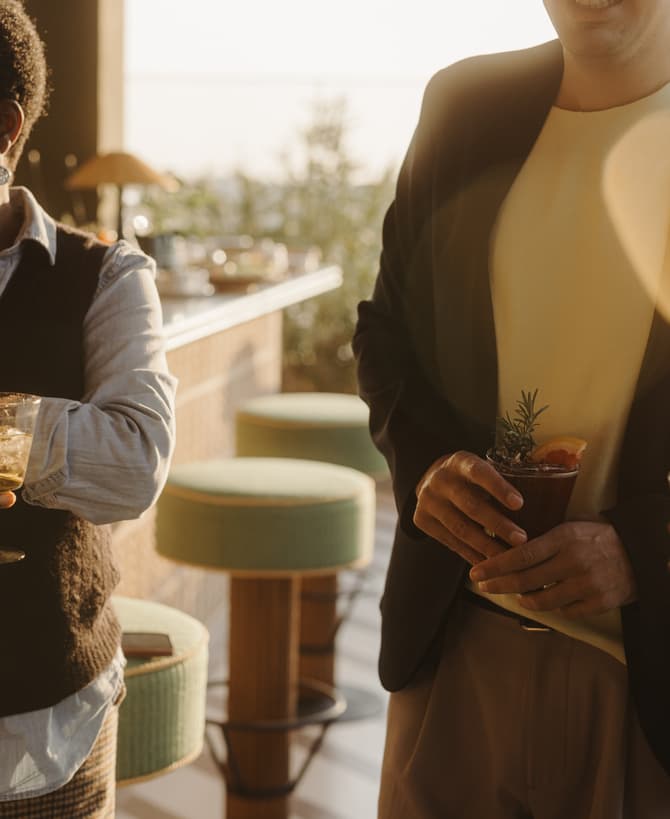 People socialize while holding cocktails, standing in a warmly lit outdoor lounge area with bar stools and plants.