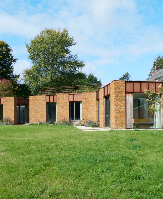 Rectangular, single-story house with stone and wood exterior, large glass doors, and a flat roof. It's set in a grassy yard with trees and adjacent traditional houses in the background.