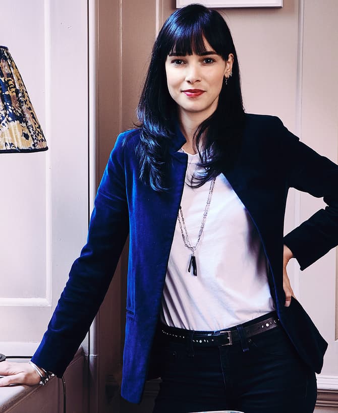 A woman in a white shirt and blue blazer stands confidently with one hand on her hip, indoors by a table with a decorative lamp and framed artwork on the walls.