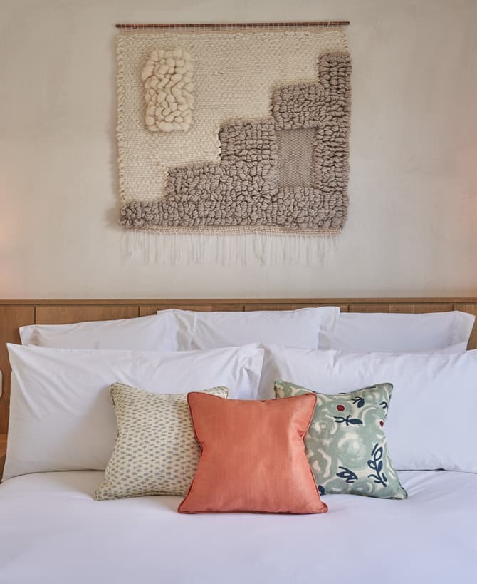 Bed adorned with white linens and three colorful pillows; flanked by wooden nightstands with lamps, phone, and clock; backdrop of woven wall hanging and framed artwork in cozy, warmly-lit bedroom.