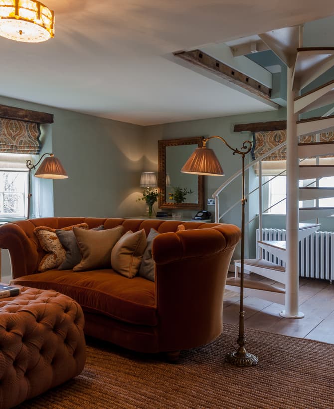 A cozy, well-lit living room with a brown tufted ottoman and matching sofa, a spiral staircase, two floor lamps, and windows with patterned blinds. A drink cart and a rug complete the setting.