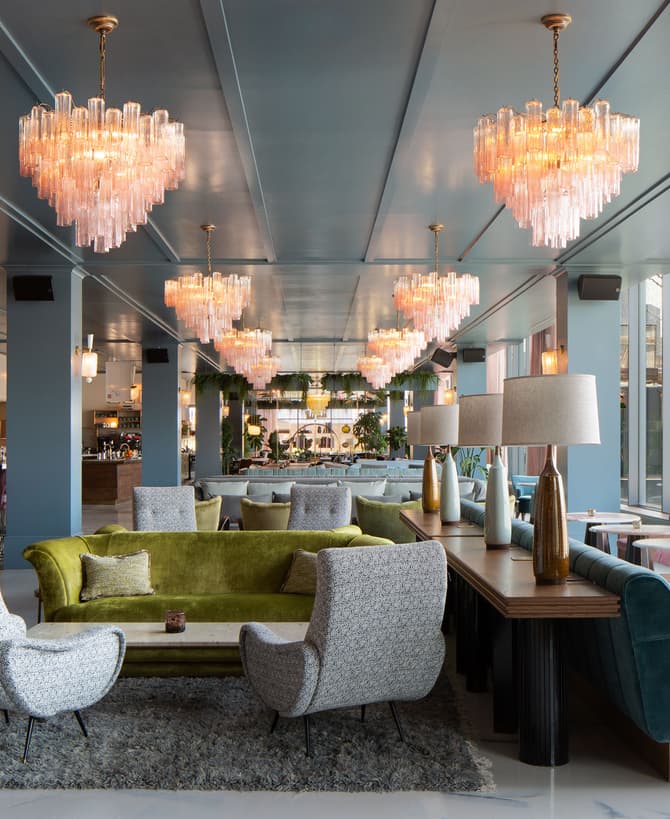 Chandeliers illuminate a modern lounge with plush seating, including a green sofa and patterned chairs, set on a carpeted floor; the space features large windows and a bar area with high stools.