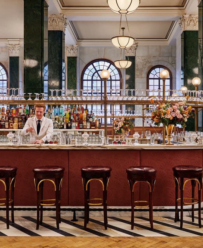 Bartender mixing drinks behind a marble counter, adorned with flower arrangements, in a luxurious bar featuring high ceilings, arched windows, hanging globe lights, and bar stools with dark cushions.