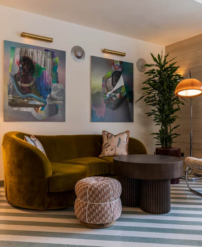 Mid-century modern living room featuring a green velvet sofa with decorative pillows, abstract wall art, a geometric table lamp, a striped ottoman, a round coffee table, a potted plant, and a unique lounge chair.