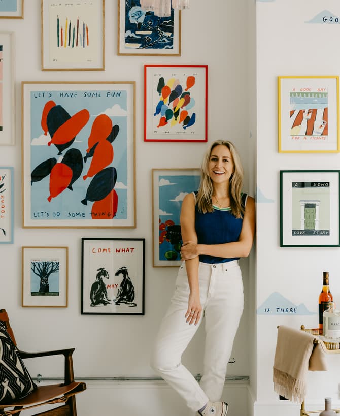 Woman leaning casually against a wall, smiling, surrounded by framed colorful artwork in a brightly lit room with red-striped wallpaper on the left.