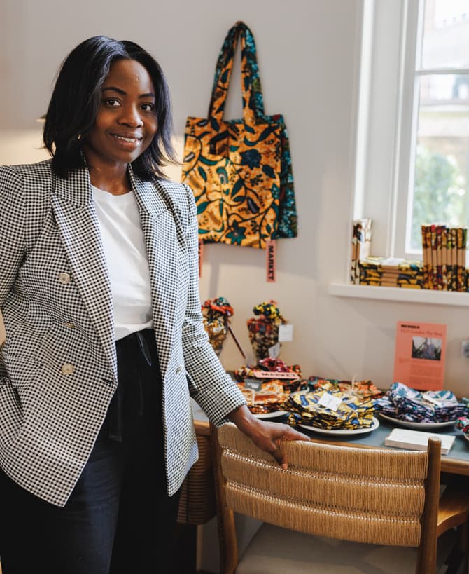 A person wearing a checkered blazer stands beside a table displaying colorful fabrics. The setting is a well-lit room with shelves containing various items and a window in the background.