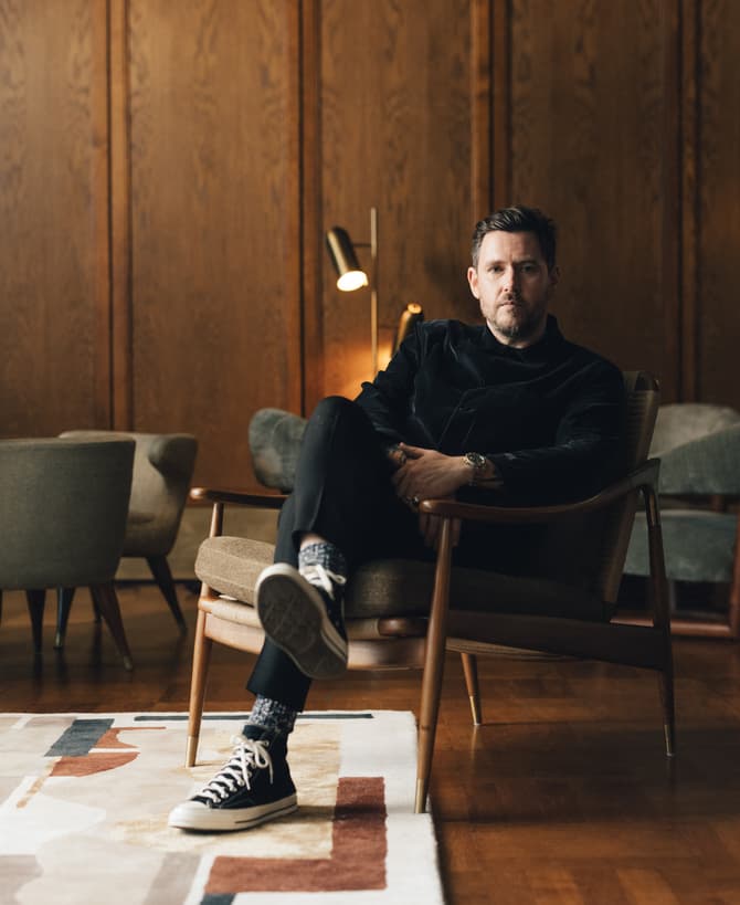 A man sits cross-legged in an armchair, wearing casual clothing and sneakers, in a mid-century modern wood-paneled room with stylish furniture and a patterned rug.