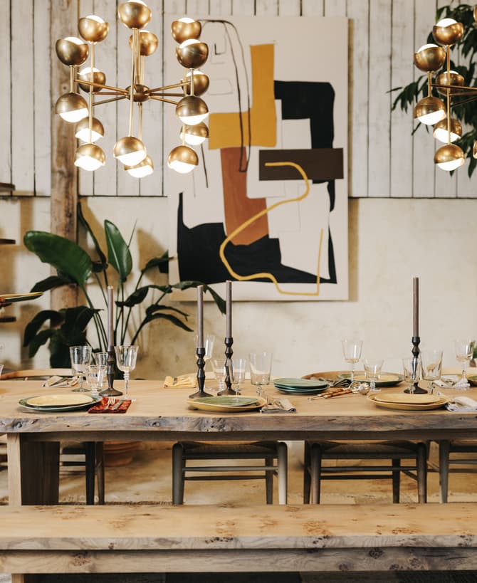 A woman in a light blue sweater is setting a dining table with plates and glassware in a rustic, well-decorated room with plants, shelves, abstract art, and gold chandeliers.