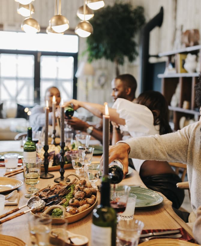 People are sitting around a wooden dining table, eating, drinking, and conversing. Two individuals are pouring wine, surrounded by plates of food, candles, and modern lighting in a cozy, well-decorated room.