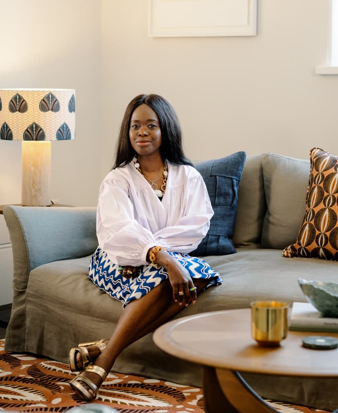 Woman sitting on a grey sofa in a cozy living room, wearing a white blouse and blue patterned skirt. The room features a patterned lampshade, decorative pillows, and a wooden coffee table with books and a candle.
