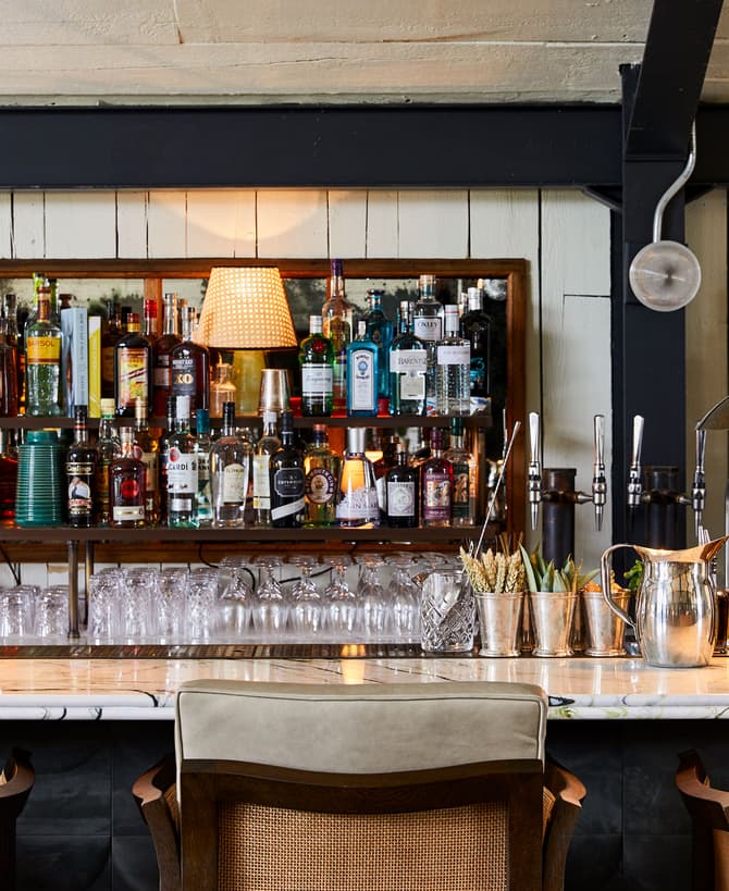 A well-stocked bar, featuring rows of liquor bottles and glassware, is lined with plush bar stools and equipped with silver utensils and lamps, creating an inviting atmosphere for patrons.