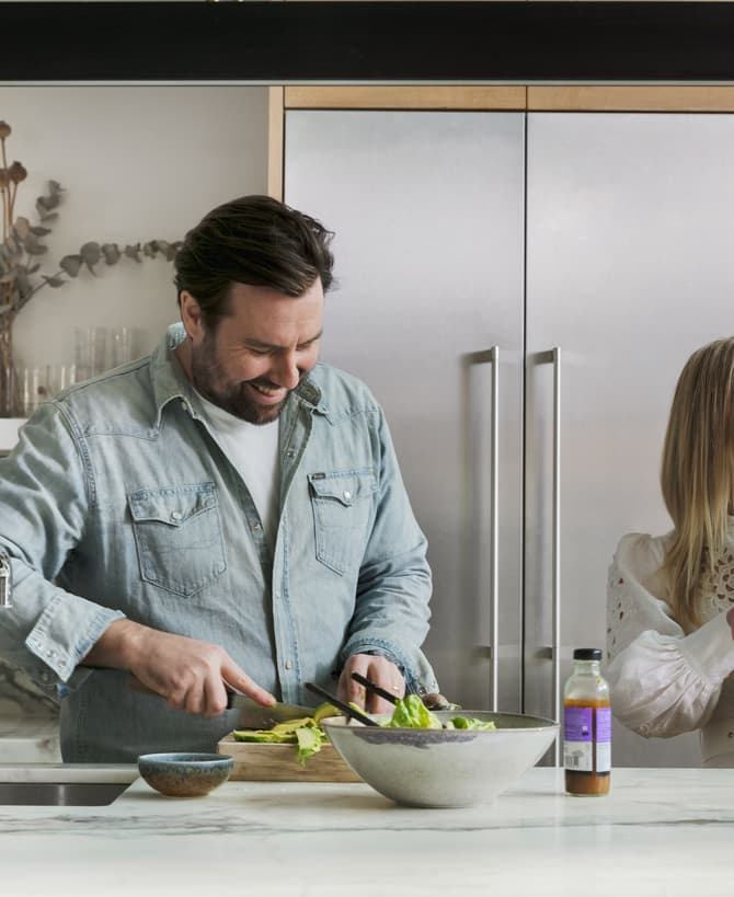 Man preparing a salad in a modern kitchen while smiling, with a woman holding a baby nearby. The kitchen features a marble countertop, sink, and stainless steel appliances.