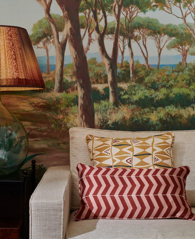 Lampshade on a glass lamp sits on a side table beside a beige sofa with patterned cushions, against a mural of a vibrant forest scene.