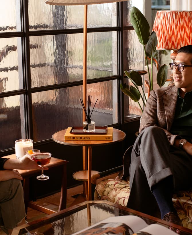 Two people sit in a cozy, well-lit room with patterned vintage decor, engaging in conversation. One holds a drink; a book titled "DUNHUANG" is on the table between them.