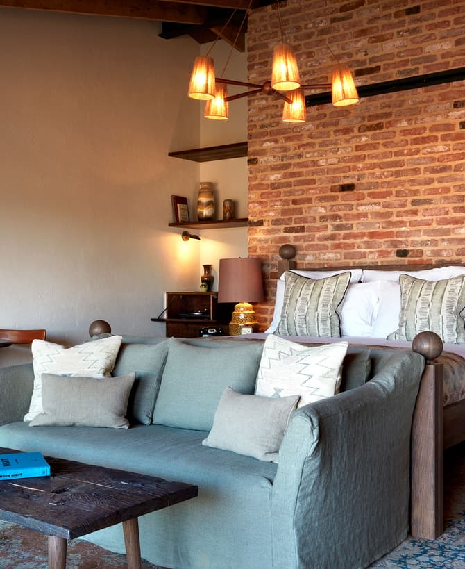 A well-lit bedroom with a brick accent wall features a wooden bed with white bedding and decorative pillows, a green sofa, wooden coffee table with a blue book, floor lamp, and small round table with chairs.