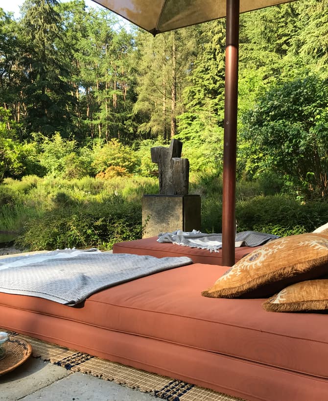 A shaded outdoor seating area features an orange cushioned daybed with pillows, a blanket, and a tea set, surrounded by lush, green vegetation and trees.