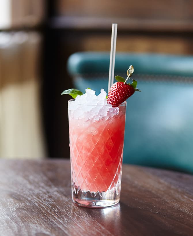 A red cocktail with ice, a straw, and a garnished strawberry sits on a wooden table in front of a blurred, elegant indoor setting.