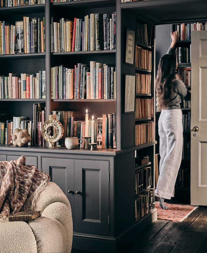 A woman in white pants and a gray top reaches for a book on tall, built-in bookshelves with numerous books; a beige, textured armchair and decor are in front.