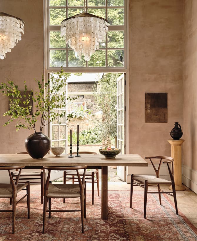 Dining table with chairs and a large vase with branches, positioned below two chandeliers, in a bright room featuring large floor-to-ceiling paneled windows and walls with artwork, providing a view to a garden.