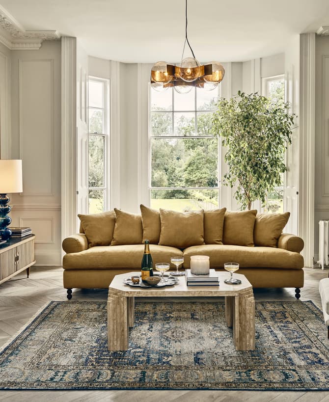 A mustard-yellow sofa with five cushions sits in a sophisticated living room with large windows, a leafy indoor plant, and elegant decor including a patterned rug, a modern coffee table, and wall art.