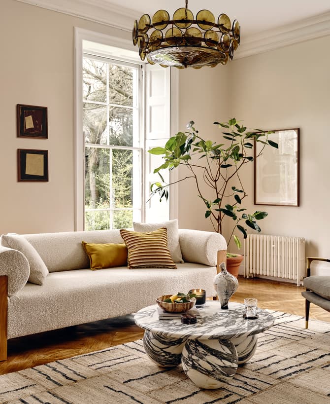 A cozy living room features a white textured sofa with yellow cushions, a marble coffee table, a gray armchair, and a large potted plant, surrounded by tall windows overlooking a green outdoor scene.
